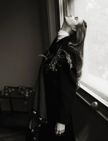 Monochrome portrait of a woman in a dark coat standing by a window with decorative accents. Interior setting with natural light.