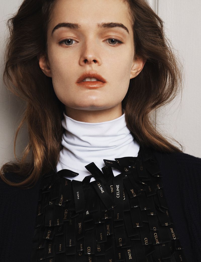 Close-up fashion shot of a fair-skinned woman wearing a white turtleneck, black jacket, and ribbon-embellished top with 'CELINE' text.