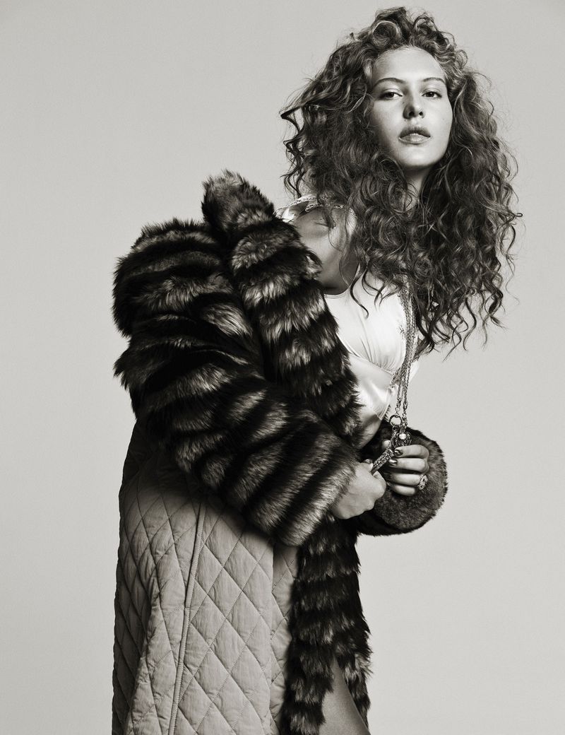 Black-and-white portrait of a woman with curly hair wearing a fur coat and long quilted skirt in a studio setting.