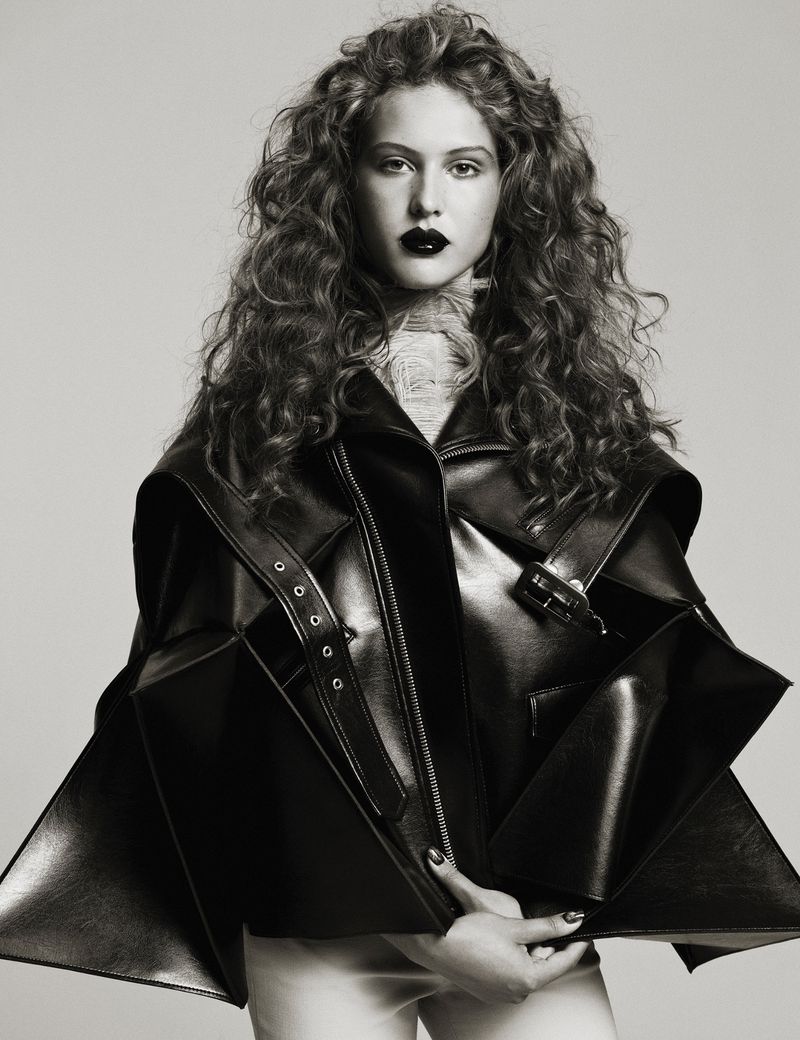 A young adult woman with curly hair and dark lipstick poses indoors wearing a black leather jacket in a studio shot.