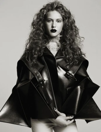 A young adult woman with curly hair and dark lipstick poses indoors wearing a black leather jacket in a studio shot.
