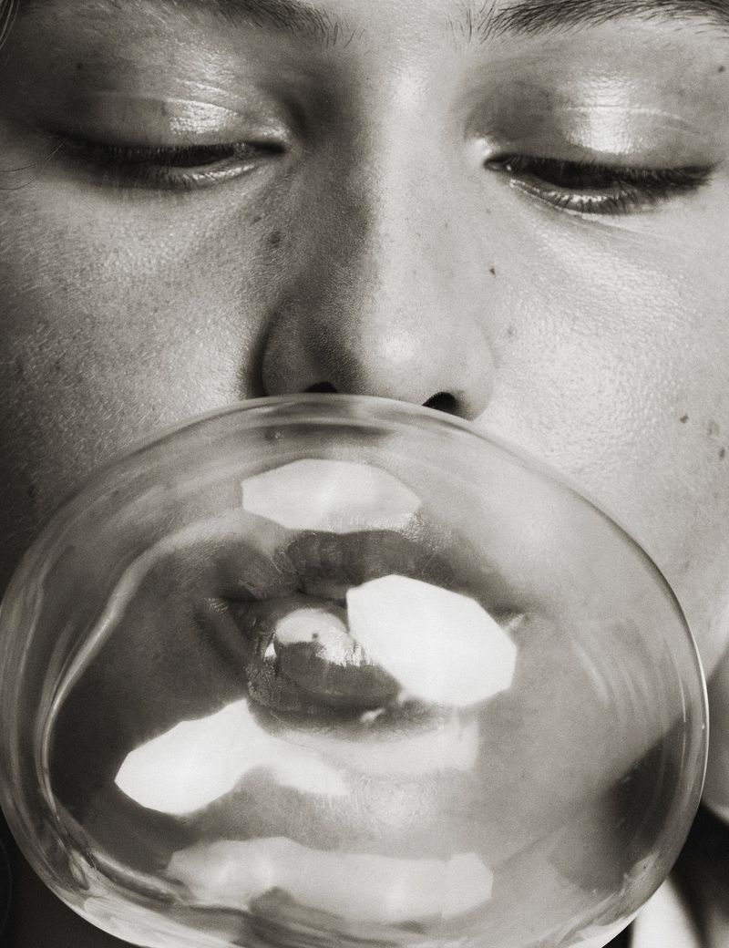 Close-up black and white portrait of a young adult blowing a bubblegum bubble, eyes closed, minimal lighting, neutral expression.