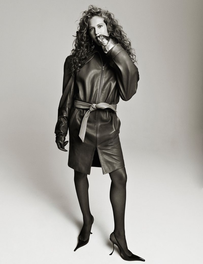 Fashion shot of a curly-haired woman in a leather dress and heels, striking a pose in a photo studio with a light grey background.