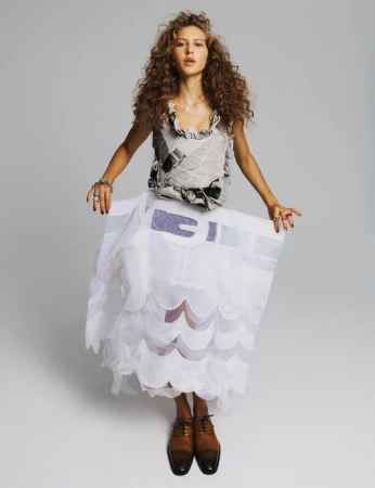 Full-length shot of a woman with curly hair wearing a newsprint-patterned top and layered white skirt, posing in a light studio.
