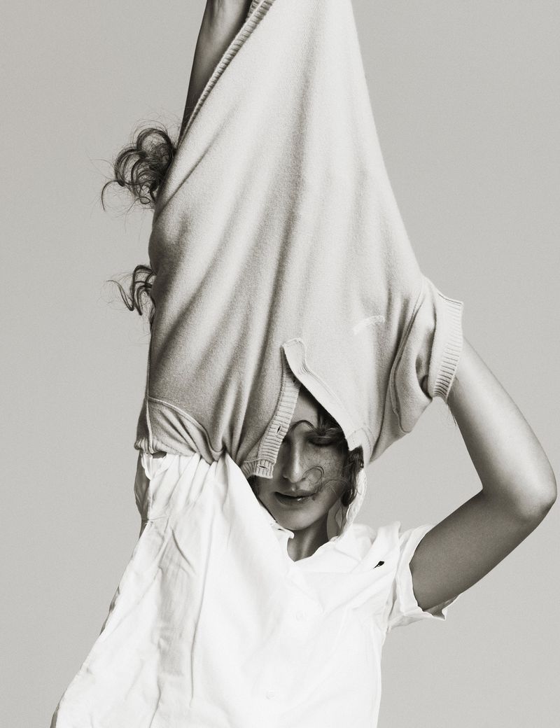 Monochromatic studio shot of a woman with curly hair indoors, wearing a white t-shirt, with a sweater pulled over her head.