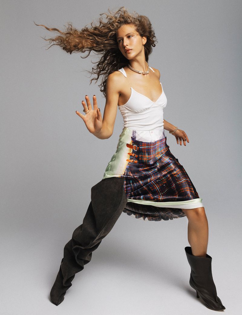 A fair-skinned female fashion model poses dynamically in a white tank top and unique skirt and gray boot combination against a light gray backdrop.