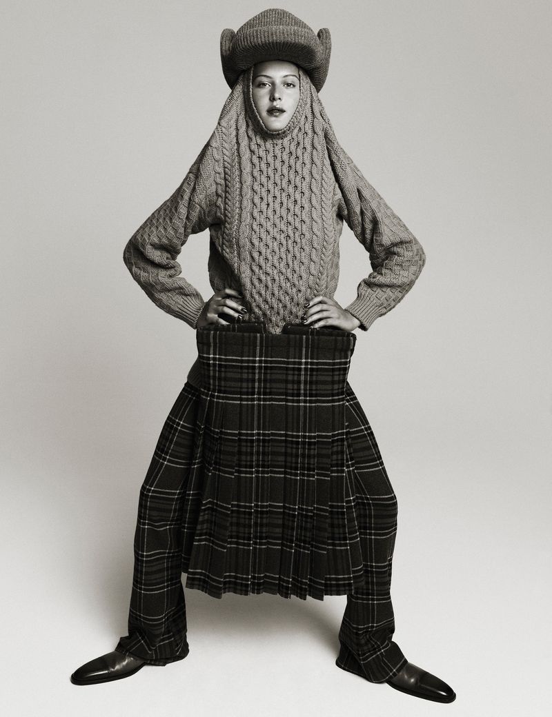 A person wearing a gray, cable-knit sweater and matching hat with a plaid kilt poses in front of a neutral background.