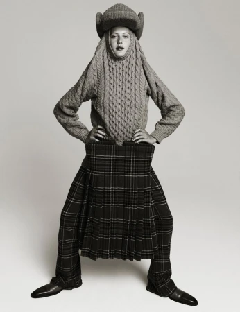 A person wearing a gray, cable-knit sweater and matching hat with a plaid kilt poses in front of a neutral background.