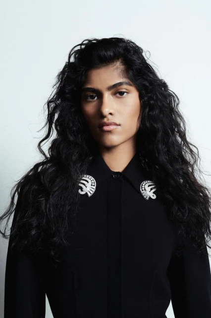 Fashion portrait of a South Asian woman with dark, curly hair wearing a black collared shirt adorned with two ornamental brooches.
