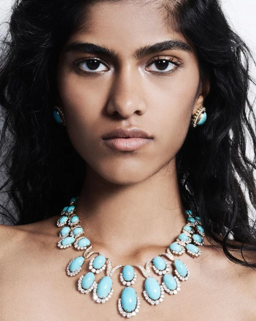 Close-up of a woman with fair skin wearing turquoise jewelry, including a necklace and earrings, against a light gray background.