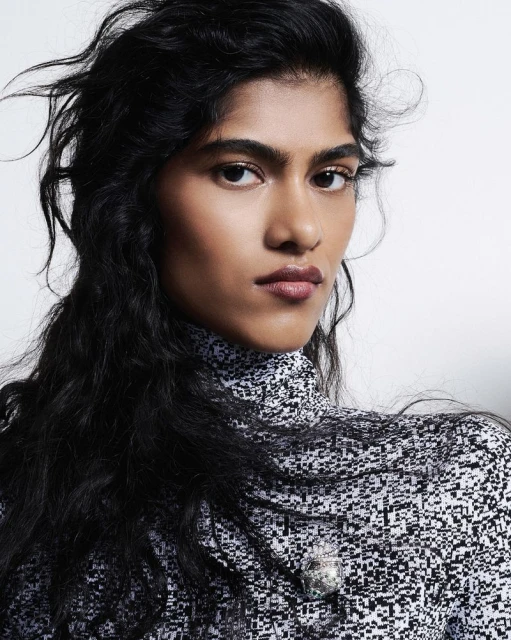 Portrait of an adult female with long, dark hair, wearing a patterned high-neck top in a studio setting with a plain background.