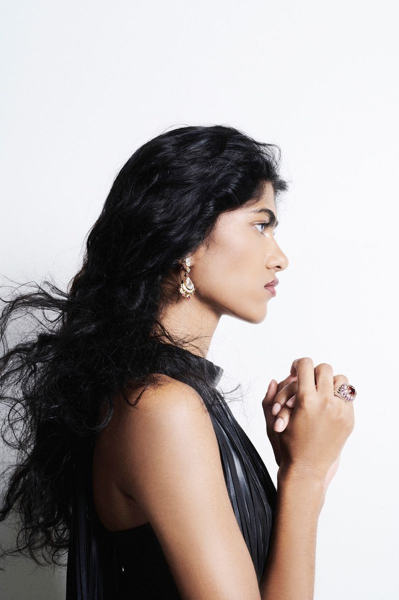 Ashley Radjarame in profile with windblown dark hair and clasped hands wearing jewelry against a white background.