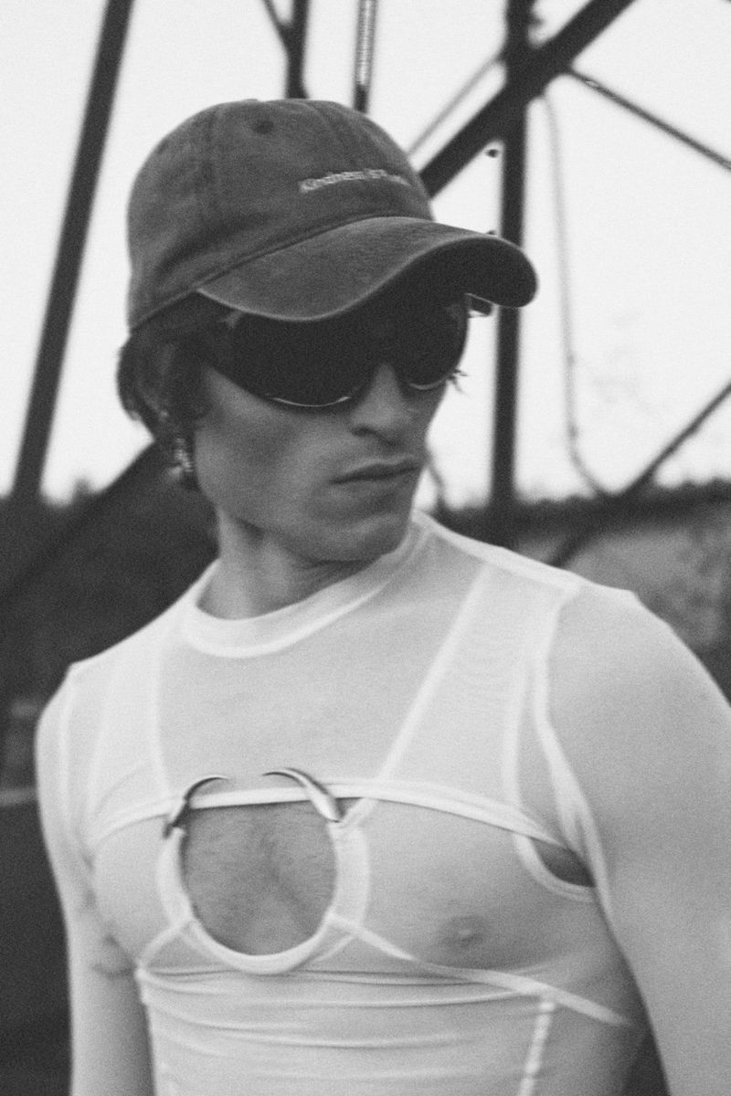 Monochrome portrait of a young man wearing a cap and sunglasses, posing against a structural background with an artistic filter.