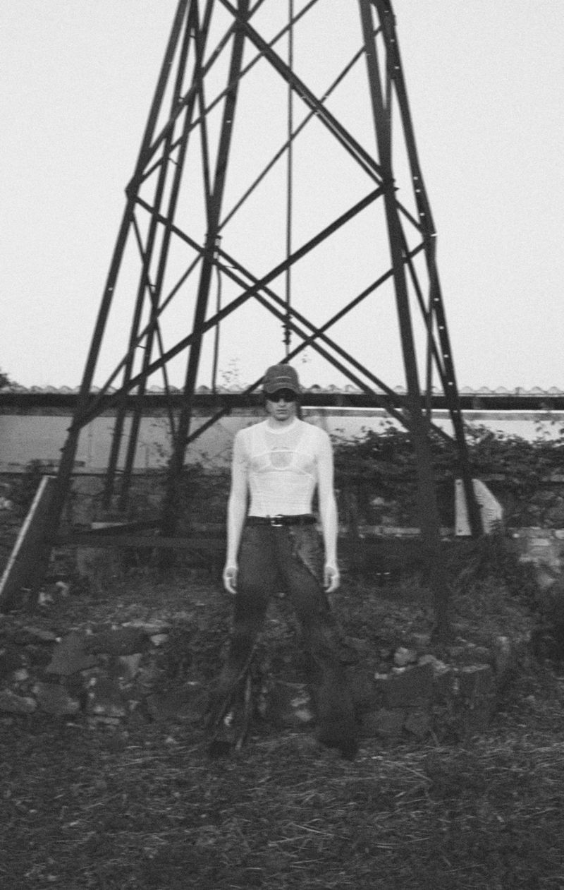 Black and white shot of a person wearing a cap, glasses, and a top standing in front of a large metal tower in a grassy area.