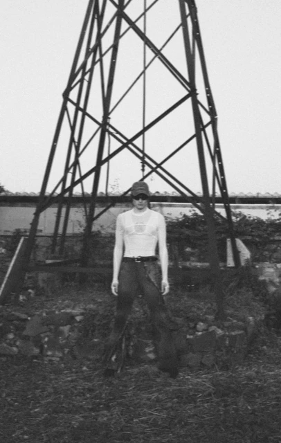 Black and white shot of a person wearing a cap, glasses, and a top standing in front of a large metal tower in a grassy area.