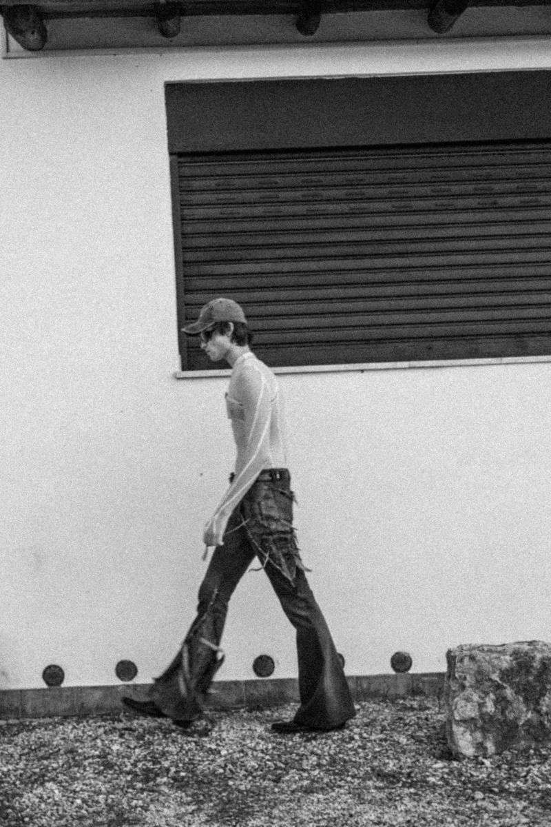 A slender adult in a baseball cap, jeans, and boots walking across a gravel surface with a light-colored building in the background.