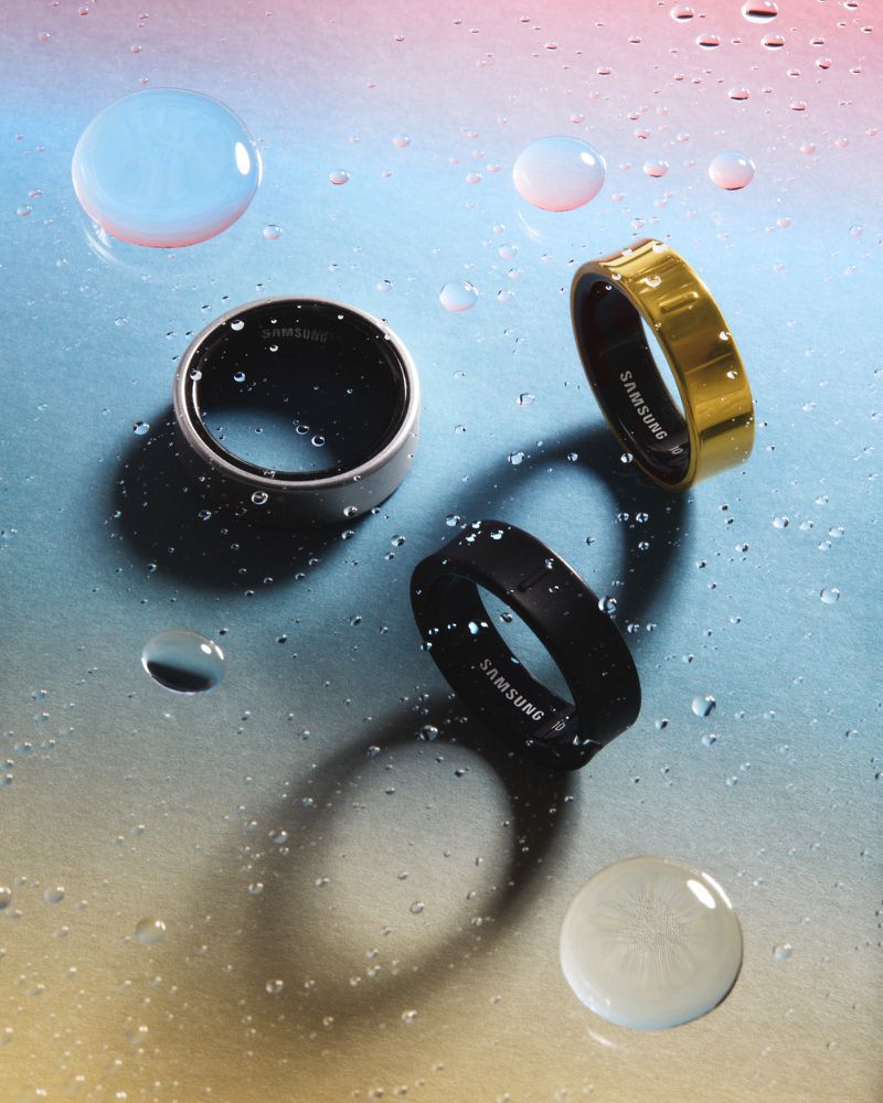 Three Samsung smart rings are displayed on a wet surface, showcasing a silver, black, and gold variant with water droplets.