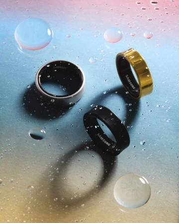 Three Samsung smart rings are displayed on a wet surface, showcasing a silver, black, and gold variant with water droplets.