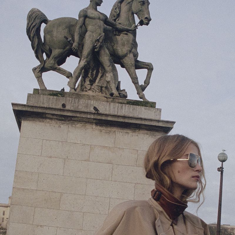 Adult female poses outdoors wearing sunglasses and a trench coat, with a large statue of a man and horse in the background on a cloudy day.