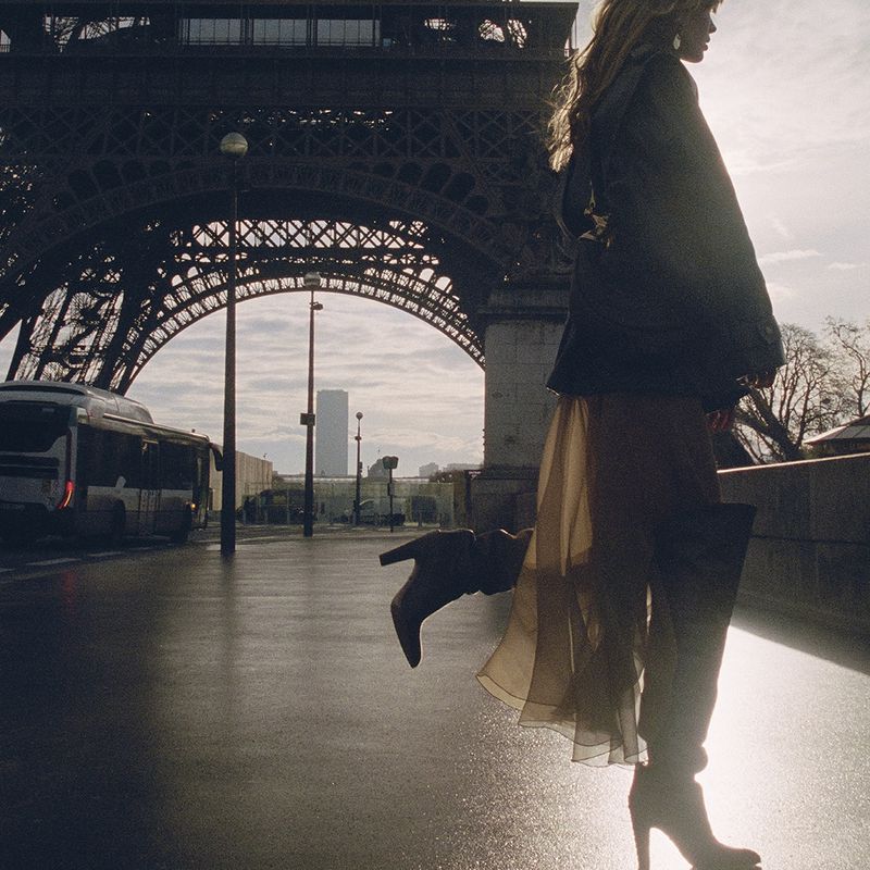 Model standing outdoors near a bridge and landmark structure wearing a dark jacket and sheer skirt with boots