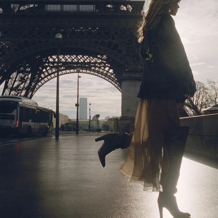 Model standing outdoors near a bridge and landmark structure wearing a dark jacket and sheer skirt with boots