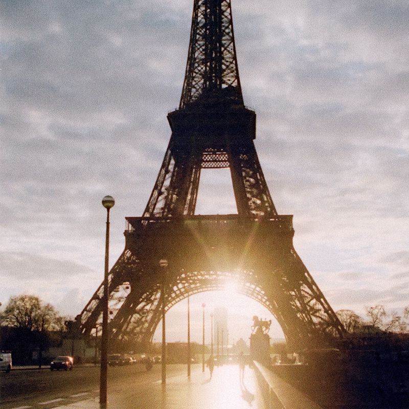 Eiffel Tower in Paris at sunrise with sun flares, street lamps, and a wet street. The sky is overcast, and the tower is silhouetted.