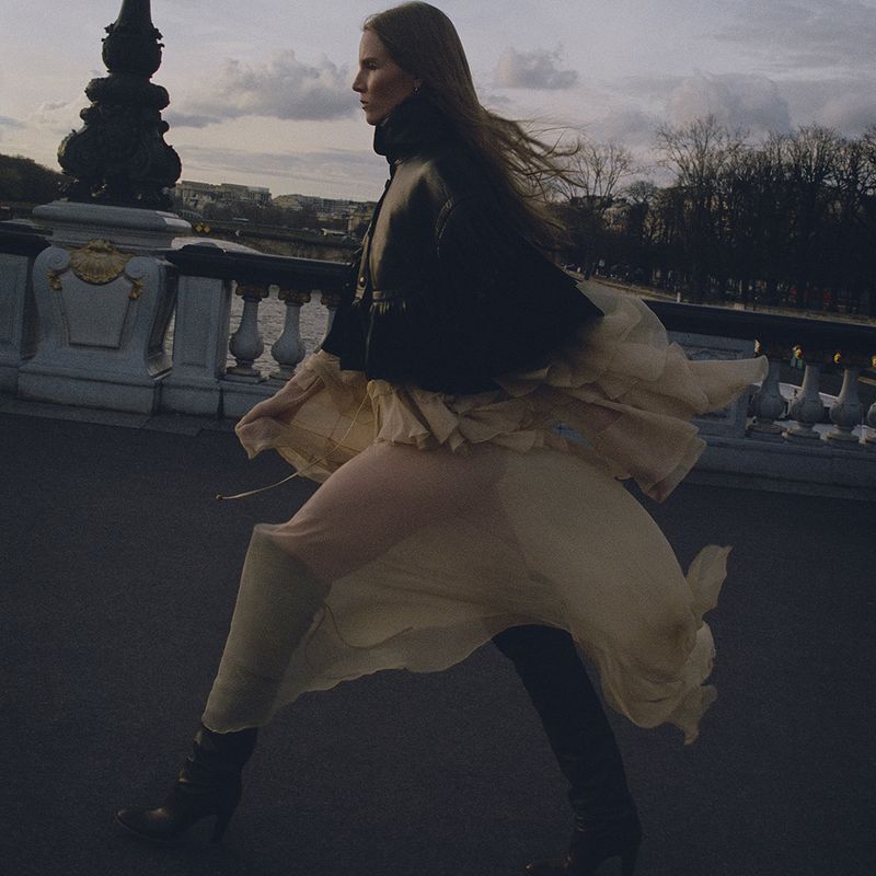 Fashionable adult female wearing a leather jacket and flowing skirt walking on a bridge with a cityscape in the background during daytime.