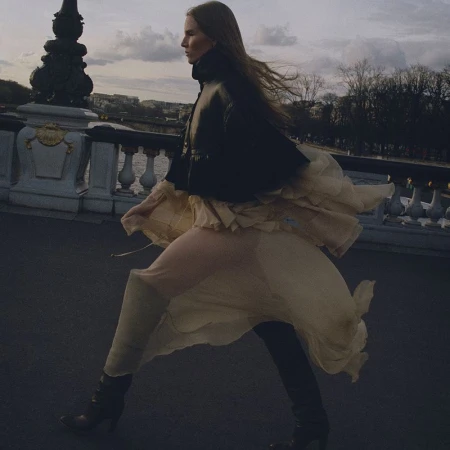 Fashionable adult female wearing a leather jacket and flowing skirt walking on a bridge with a cityscape in the background during daytime.