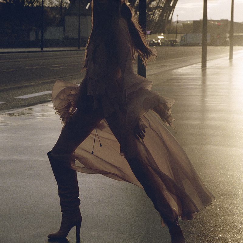 A woman wearing high-heeled boots and a flowing dress poses on a wet city street in front of the Eiffel Tower at dusk.