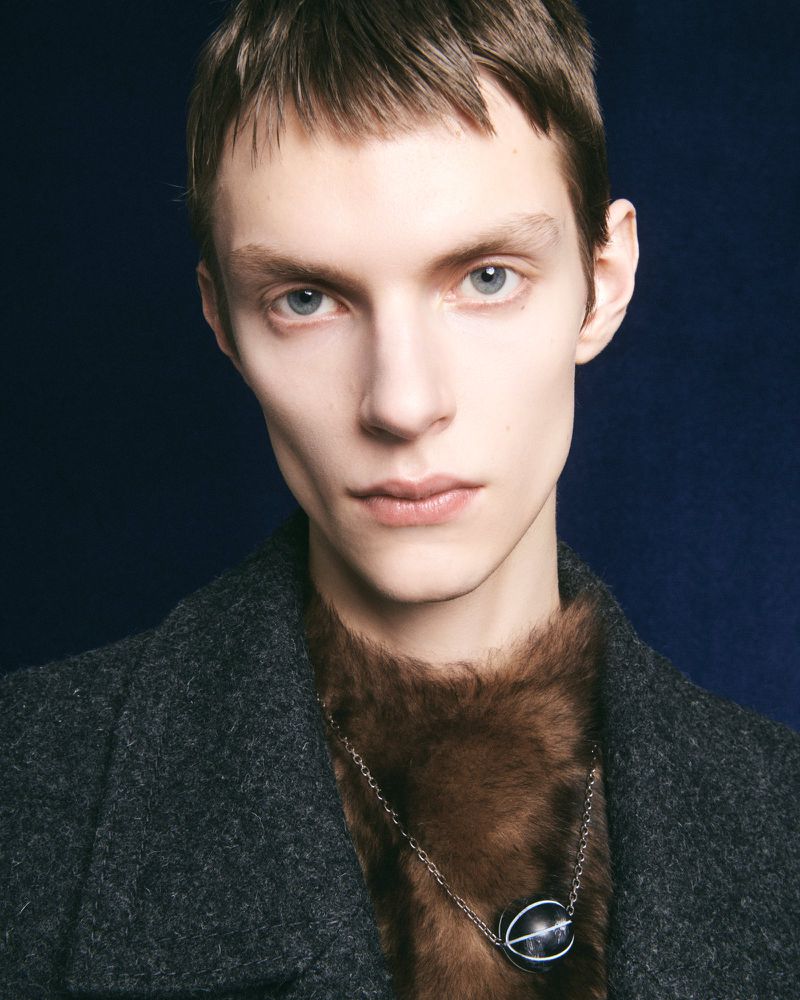 Close-up of a young adult man with light skin, wearing a grey coat and brown faux fur sweater with an oval pendant necklace.
