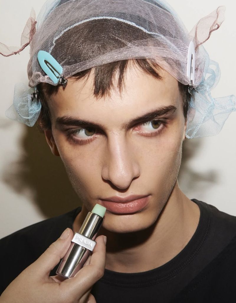 Close-up of a male model with brown hair wearing a hair net, being prepped with lip balm applied by an unseen hand. Beauty advertising.