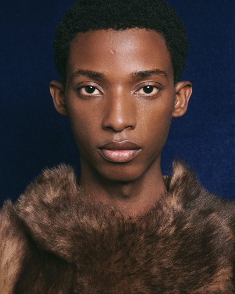 Close-up portrait of a young Black male model wearing a textured brown fur collar against a dark blue background.
