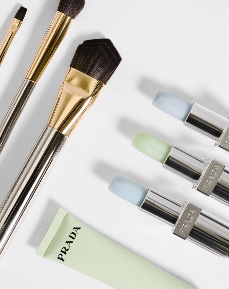 Close-up of Prada makeup brushes with silver and gold handles, and pastel color sticks displayed on a bright white surface.