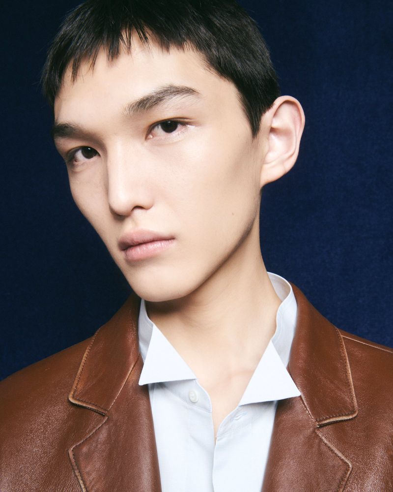 Close-up portrait of a male model wearing a brown leather jacket and light blue collared shirt against a dark textured blue background.