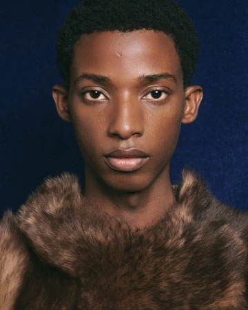 Close-up portrait of a young Black male model wearing a textured brown fur collar against a dark blue background.