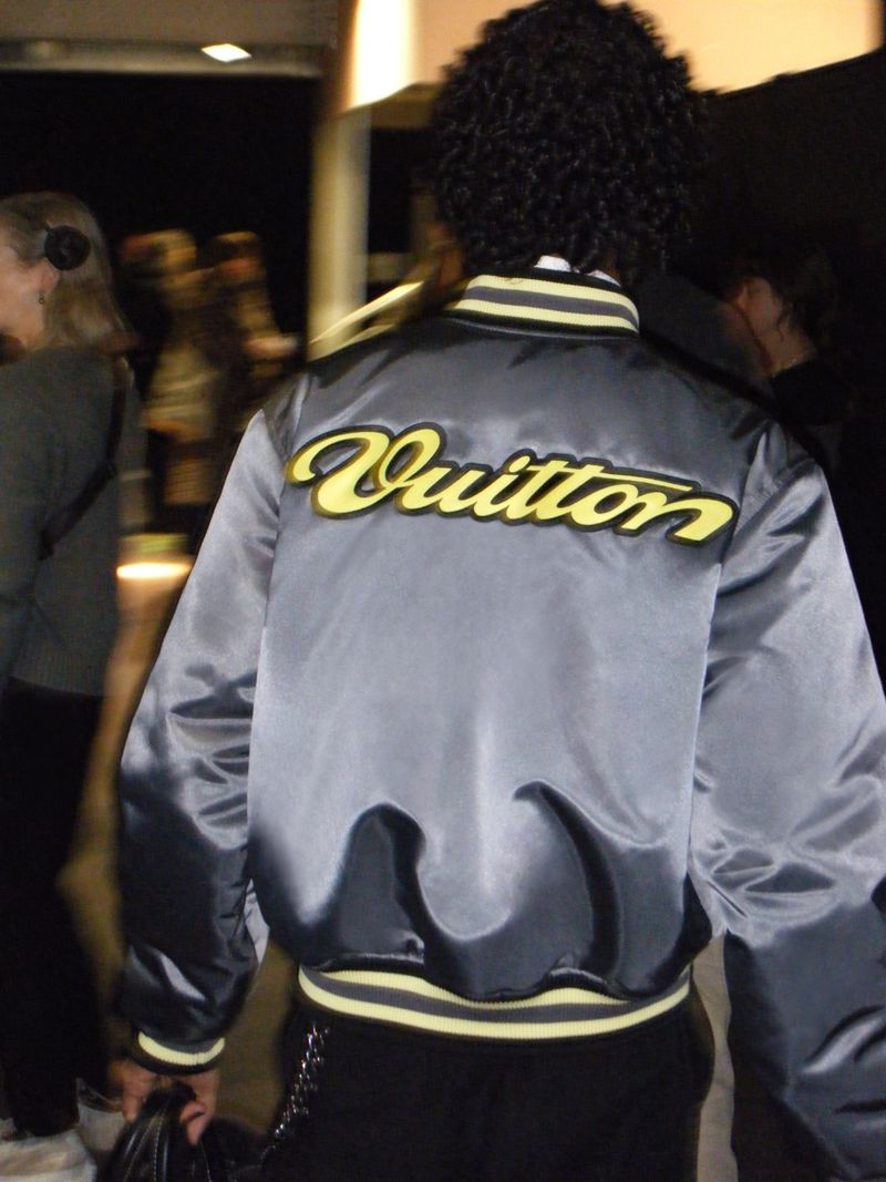 Person wearing a gray bomber jacket with yellow 'Vuitton' lettering, standing in an indoor setting with blurred figures in the background.