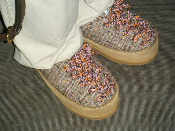 Close-up of beige slippers with textured design, worn with light-colored pants and a brown belt, set against a dark, neutral background.