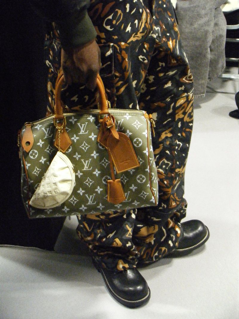 Olive Louis Vuitton Speedy bag held by a person wearing patterned pants and black shoes in an indoor setting with neutral tones.
