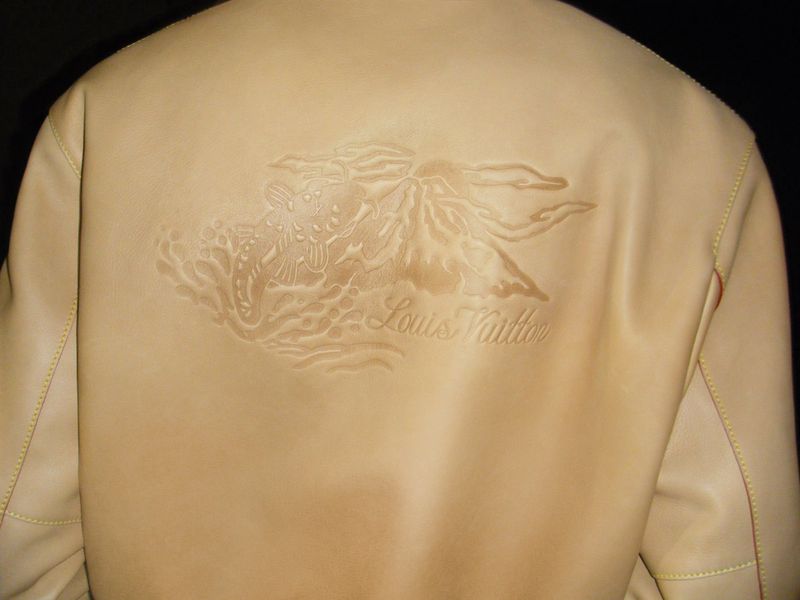 Tan leather jacket with embossed design and Louis Vuitton logo on the back, against a dark background, showcased in a studio setting.