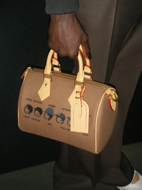 Close-up of a tan Louis Vuitton handbag with a unique graphic design, being held by a dark-skinned hand in a studio setting.