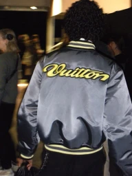 Model seen from behind wearing a glossy grey satin bomber jacket with yellow Vuitton lettering on the back
