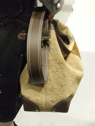 Close-up of a person wearing dark denim holding a tan monogram suede handbag by Louis Vuitton against a plain background.