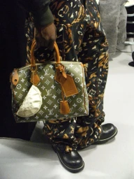 Close-up of a person holding an olive green Louis Vuitton monogram handbag wearing patterned trousers and black boots
