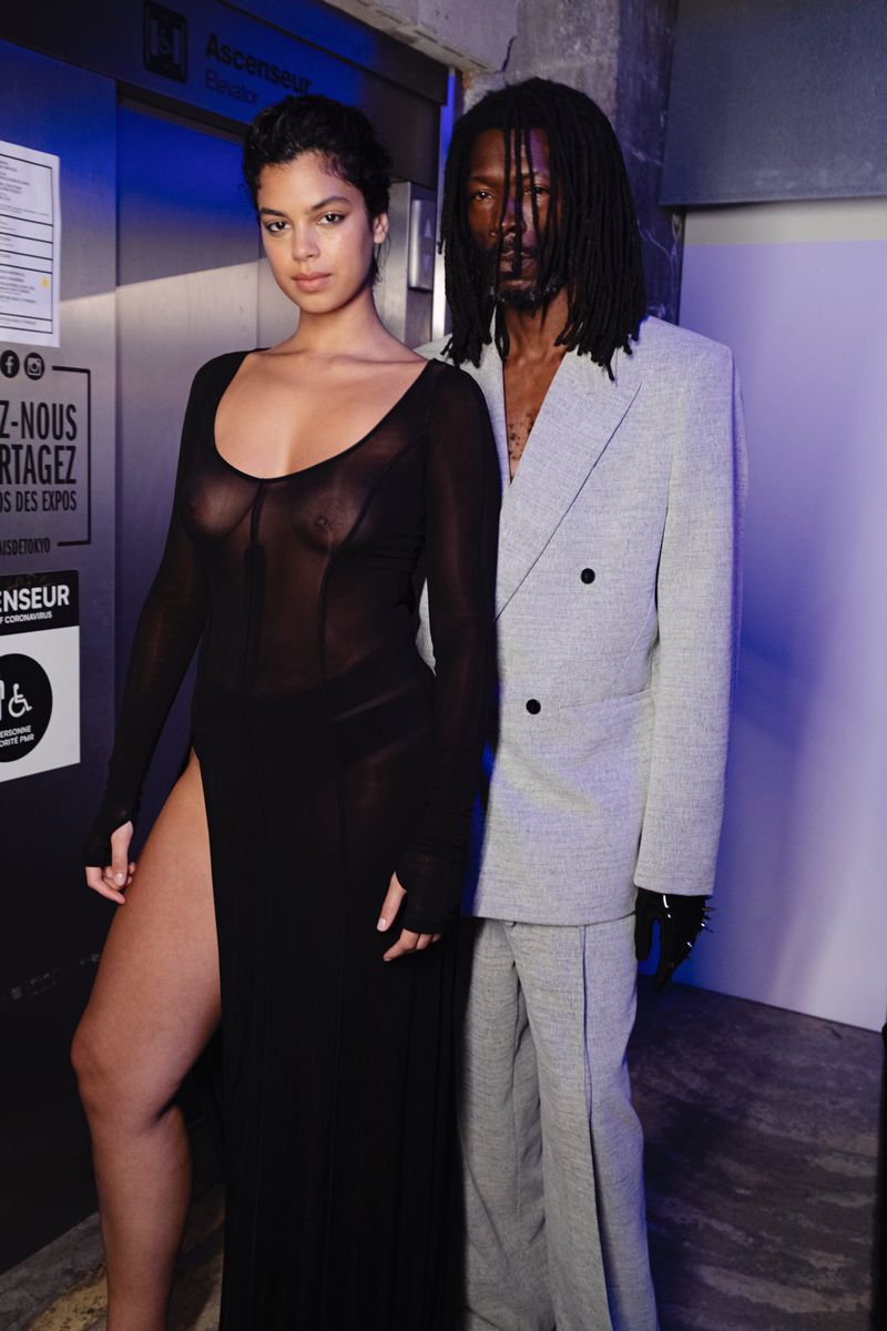 A fashionable mixed-race couple poses together in a dimly lit urban indoor setting. The woman wears a black sheer dress with a thigh-high slit.