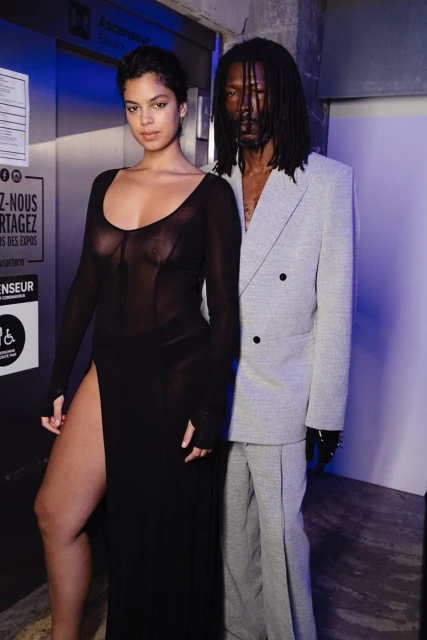 A fashionable mixed-race couple poses together in a dimly lit urban indoor setting. The woman wears a black sheer dress with a thigh-high slit.
