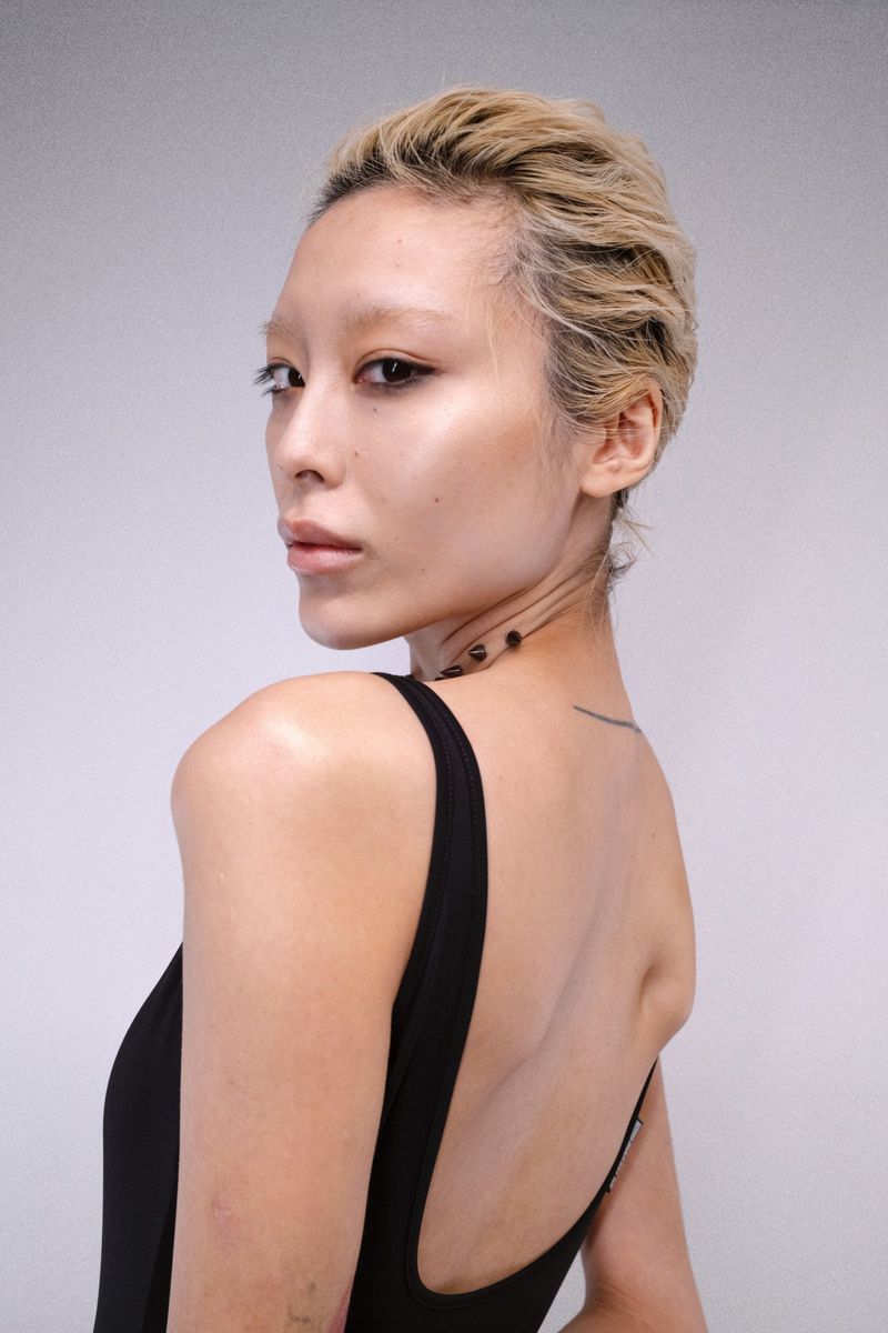 Portrait of a model with blonde hair and a black top looking back over her shoulder against a light grey wall.