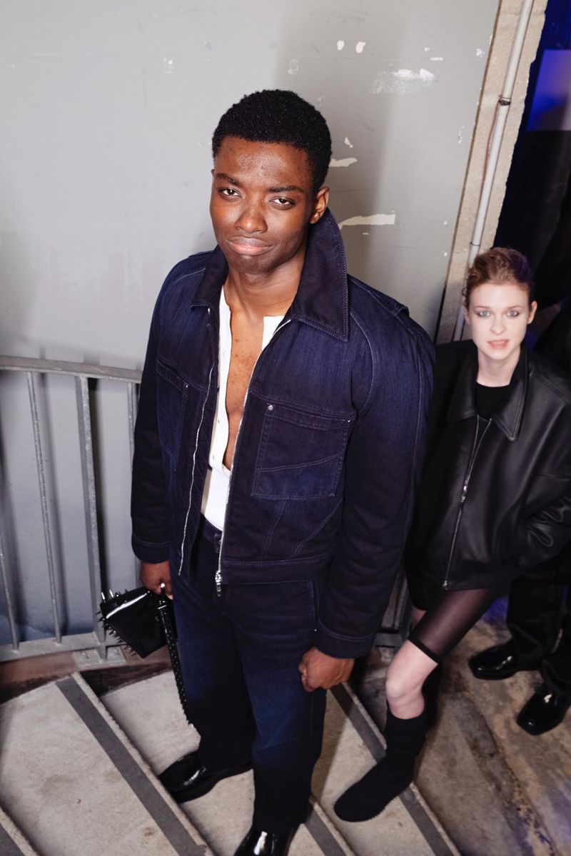 A fashionable African American man in a blue denim jacket poses on grey stairs with a stylish woman in black in the background.