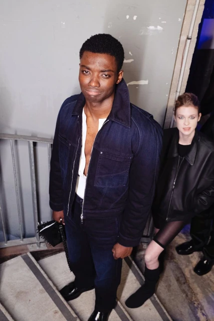 A fashionable African American man in a blue denim jacket poses on grey stairs with a stylish woman in black in the background.