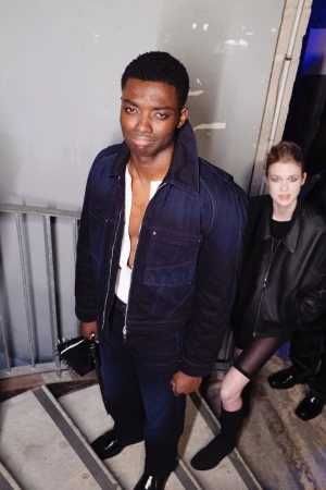 A fashionable African American man in a blue denim jacket poses on grey stairs with a stylish woman in black in the background.