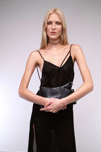 Fashion photography: Young woman poses in a black spaghetti-strap dress, holding a black clutch, against a neutral background in studio lighting.